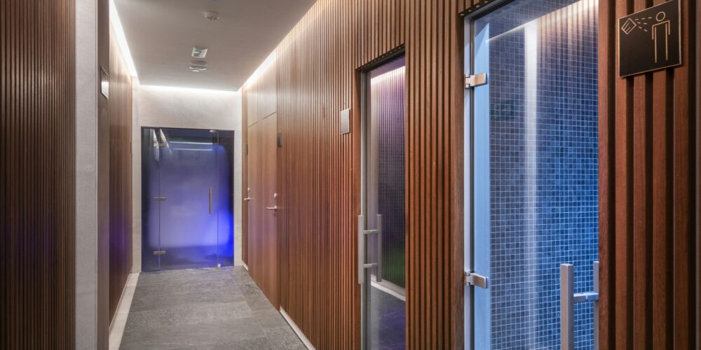 Un pasillo contemporáneo que presenta paneles de madera, listones verticales y una sutil iluminación LED. Las puertas de vidrio al final conducen a las instalaciones del spa, con una luz azul visible, y un cartel de amenidades montado cerca. El suelo está compuesto por azulejos de piedra oscura.