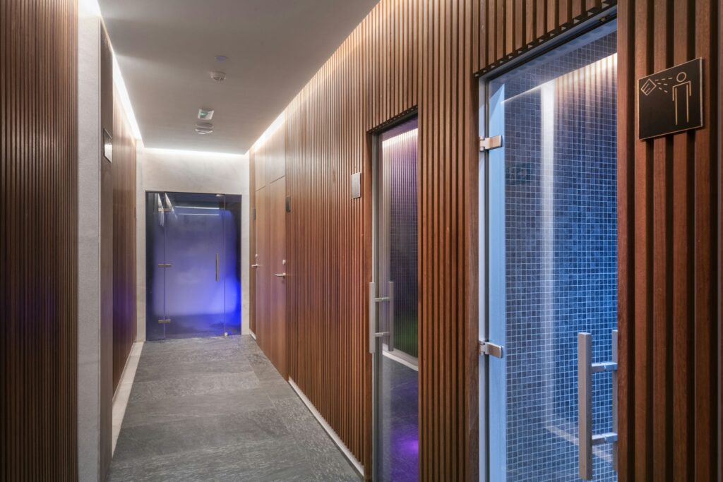Pasillo Moderno de Spa con Paneles de Madera y Iluminación LED Un pasillo contemporáneo que presenta paneles de madera, listones verticales y una sutil iluminación LED. Las puertas de vidrio al final conducen a las instalaciones del spa, con una luz azul visible, y un cartel de amenidades montado cerca. El suelo está compuesto por azulejos de piedra oscura.