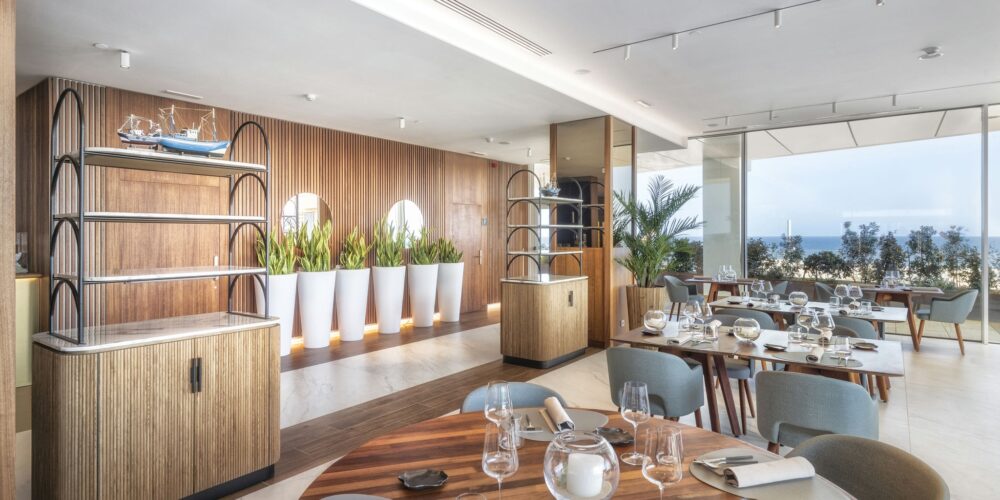Interior de restaurante moderno con grandes ventanas que dan al océano, elegantes acentos de madera, mesas circulares y rectangulares decoradas con cristalería y servilletas blancas. Incluye una unidad de estantería decorativa con un modelo de barco y grandes macetas blancas llenas de vegetación, creando una atmósfera luminosa y sofisticada para cenar.