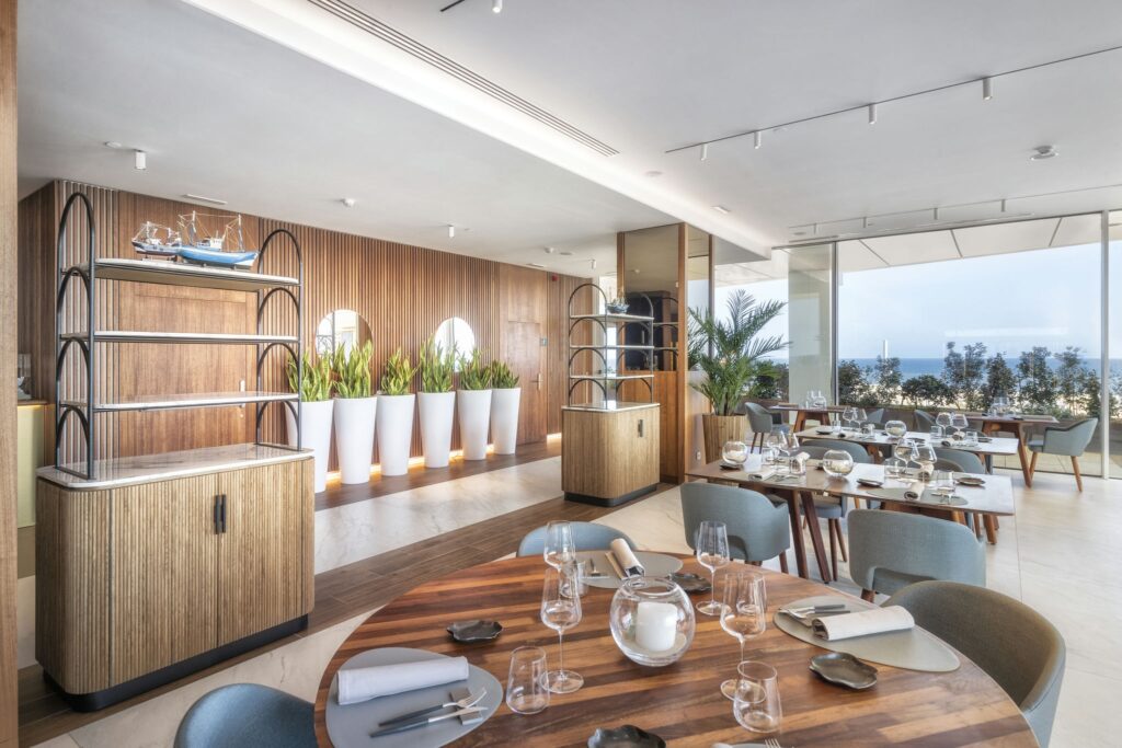 Interior de Restaurante Elegante con Vista al Océano y Acentos de Madera Interior de restaurante moderno con grandes ventanas que dan al océano, elegantes acentos de madera, mesas circulares y rectangulares decoradas con cristalería y servilletas blancas. Incluye una unidad de estantería decorativa con un modelo de barco y grandes macetas blancas llenas de vegetación, creando una atmósfera luminosa y sofisticada para cenar.