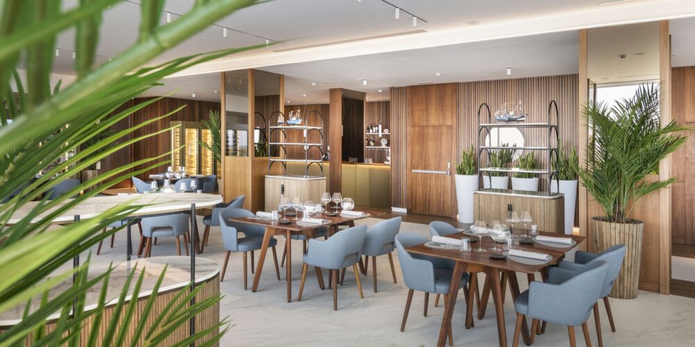 Un restaurante espacioso y contemporáneo con acentos de madera, sillas tapizadas en azul, platos blancos y cristalería decorativa. El interior es luminoso y realza la experiencia gastronómica con una exuberante vegetación y un área de bar sofisticada.