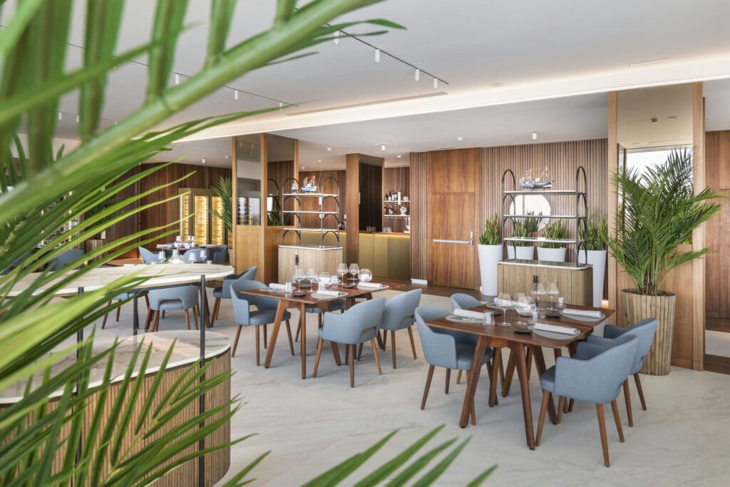 Interiores de Restaurante Estiloso y Moderno con Decoración Elegante y Accentos de Madera Un restaurante espacioso y contemporáneo con acentos de madera, sillas tapizadas en azul, platos blancos y cristalería decorativa. El interior es luminoso y realza la experiencia gastronómica con una exuberante vegetación y un área de bar sofisticada.