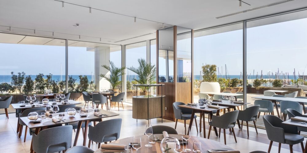 Interior estiloso de un restaurante moderno que presenta mobiliario de madera elegante, mesas de comedor dispuestas con buen gusto, vajilla, copas de vino y servilletas. Grandes ventanales de vidrio ofrecen una vista panorámica del océano y la marina, complementada por una vegetación exuberante que crea una atmósfera acogedora.