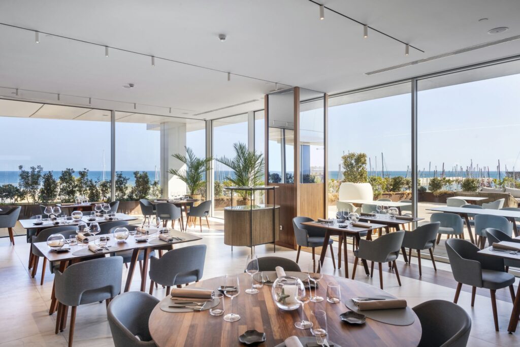 Interiores Elegantes y Modernos de Restaurante con Vista al Océano Interior estiloso de un restaurante moderno que presenta mobiliario de madera elegante, mesas de comedor dispuestas con buen gusto, vajilla, copas de vino y servilletas. Grandes ventanales de vidrio ofrecen una vista panorámica del océano y la marina, complementada por una vegetación exuberante que crea una atmósfera acogedora.