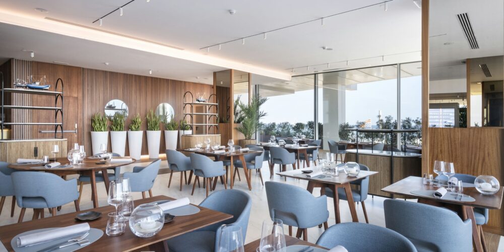 Interiores de restaurante elegante con mesas de madera, utensilios y copas de mesa dispuestos ordenadamente, sillas azules, una pared de madera con paneles verticales, estanterías, espejos circulares y exuberantes plantas verdes en macetas blancas, iluminadas por la luz natural de grandes ventanas.
