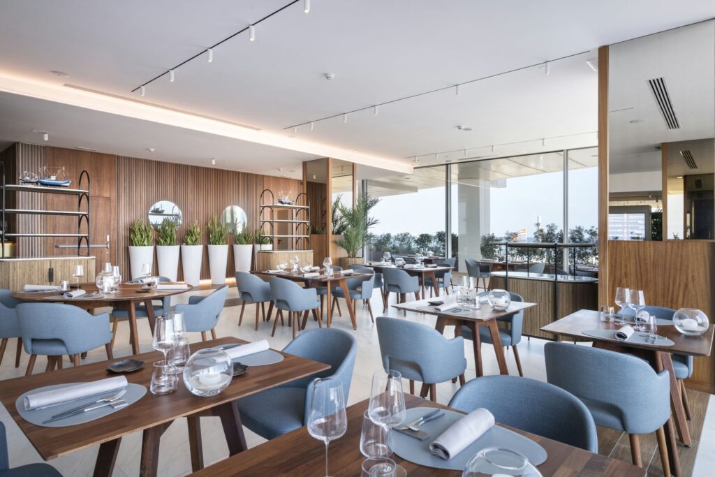 Interior Elegante y Moderno de Restaurante con Diseño Minimalista Interiores de restaurante elegante con mesas de madera, utensilios y copas de mesa dispuestos ordenadamente, sillas azules, una pared de madera con paneles verticales, estanterías, espejos circulares y exuberantes plantas verdes en macetas blancas, iluminadas por la luz natural de grandes ventanas.