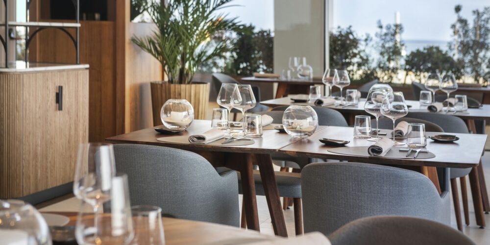 Hotel Marina Badalona: Interior de restaurante con estilo que exhibe mesas de comedor de madera elegantemente dispuestas con cristalería, incluyendo copas de vino y vasos. Servilletas bien enrolladas y pequeños platos están organizados en cada mesa, creando una atmósfera de comedor sofisticada. La luz natural inunda el espacio a través de grandes ventanales, ofreciendo una vista de la vegetación exuberante del exterior. La estética moderna se complementa con sillas tapizadas en gris y una decoración minimalista, invitando a los huéspedes a disfrutar de una agradable experiencia gastronómica.