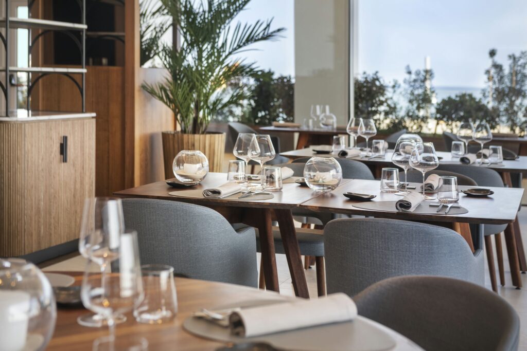 Hotel Marina Badalona: Interior de restaurante con estilo que exhibe mesas de comedor de madera elegantemente dispuestas con cristalería, incluyendo copas de vino y vasos. Servilletas bien enrolladas y pequeños platos están organizados en cada mesa, creando una atmósfera de comedor sofisticada. La luz natural inunda el espacio a través de grandes ventanales, ofreciendo una vista de la vegetación exuberante del exterior. La estética moderna se complementa con sillas tapizadas en gris y una decoración minimalista, invitando a los huéspedes a disfrutar de una agradable experiencia gastronómica.