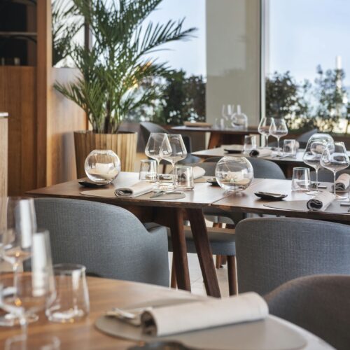 Hotel Marina Badalona: Interior de restaurante con estilo que exhibe mesas de comedor de madera elegantemente dispuestas con cristalería, incluyendo copas de vino y vasos. Servilletas bien enrolladas y pequeños platos están organizados en cada mesa, creando una atmósfera de comedor sofisticada. La luz natural inunda el espacio a través de grandes ventanales, ofreciendo una vista de la vegetación exuberante del exterior. La estética moderna se complementa con sillas tapizadas en gris y una decoración minimalista, invitando a los huéspedes a disfrutar de una agradable experiencia gastronómica.