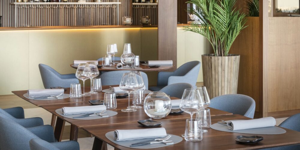 Un interior de restaurante moderno que muestra un área de comedor bellamente dispuesta con elegantes mesas de madera, vajilla estilosa y suaves sillas azules, complementado por un bar en el fondo y una planta alta enmacetada.