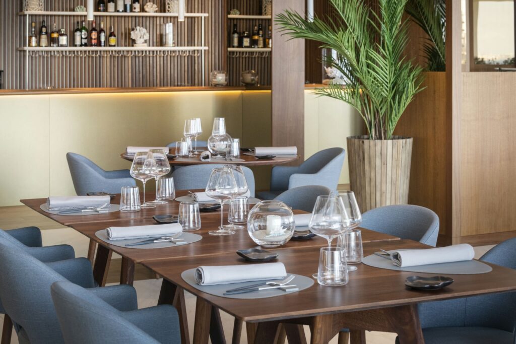 Interior de Restaurante Moderno y Elegante con Configuración de Comedor Estilosa Un interior de restaurante moderno que muestra un área de comedor bellamente dispuesta con elegantes mesas de madera, vajilla estilosa y suaves sillas azules, complementado por un bar en el fondo y una planta alta enmacetada.