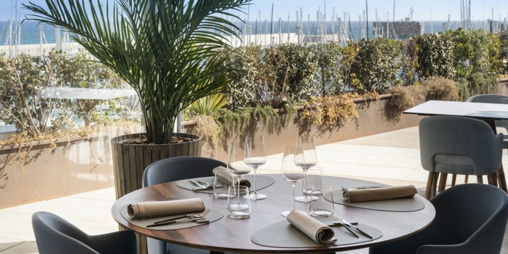 Una mesa de comedor redonda de madera bellamente dispuesta para múltiples invitados, con elegantes cristales y servilletas perfectamente dobladas, rodeada de asientos modernos. Al fondo, una gran ventana presenta una vista de exuberante vegetación y un puerto lleno de veleros bajo un brillante cielo azul, complementada por una alta planta de palma para un toque natural.