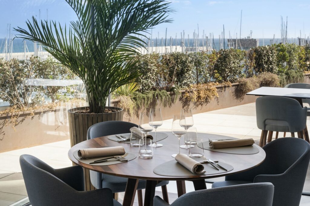Configuración de Mesa de Comedor Elegante con Vista Escénica Una mesa de comedor redonda de madera bellamente dispuesta para múltiples invitados, con elegantes cristales y servilletas perfectamente dobladas, rodeada de asientos modernos. Al fondo, una gran ventana presenta una vista de exuberante vegetación y un puerto lleno de veleros bajo un brillante cielo azul, complementada por una alta planta de palma para un toque natural.