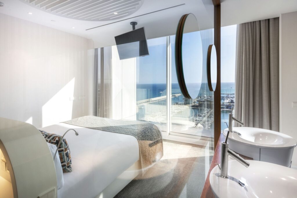 Habitación de hotel costera moderna con vista al océano Amplia habitación de hotel moderna con una cama grande con almohadas decorativas, baño elegante con dos lavabos y grandes ventanales que ofrecen una vista impresionante del océano y el paisaje costero.