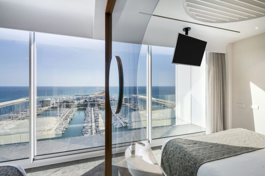 Habitación de hotel moderna y lujosa con impresionantes vistas de la marina y el océano Habitación de hotel contemporánea con grandes ventanas de vidrio, ropa de cama gris y vistas a la marina, resaltando la esencia de lujo y relajación.