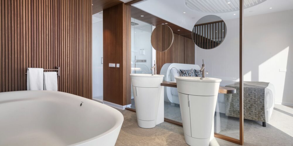 Un baño moderno con una bañera blanca independiente, dos lavabos circulares con grifos elegantes y paneles de madera cálidos. La suave luz natural ilumina el espacio, mejorando la atmósfera serena y lujosa con toallas suaves cercanas.