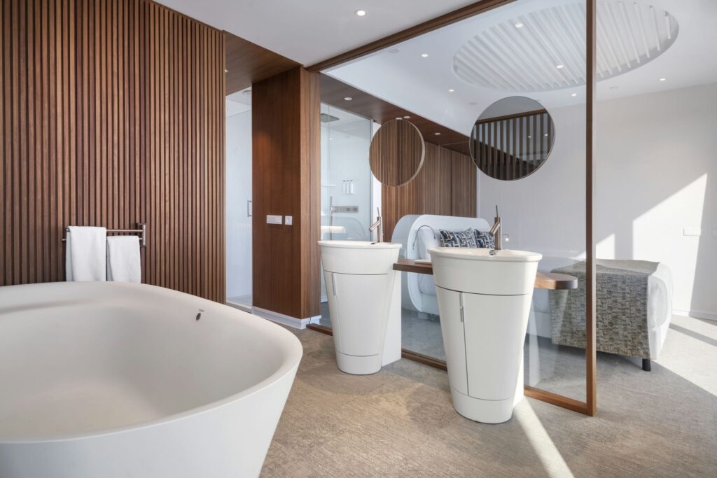 Baño de lujo contemporáneo con bañera independiente y lavabos circulares Un baño moderno con una bañera blanca independiente, dos lavabos circulares con grifos elegantes y paneles de madera cálidos. La suave luz natural ilumina el espacio, mejorando la atmósfera serena y lujosa con toallas suaves cercanas.