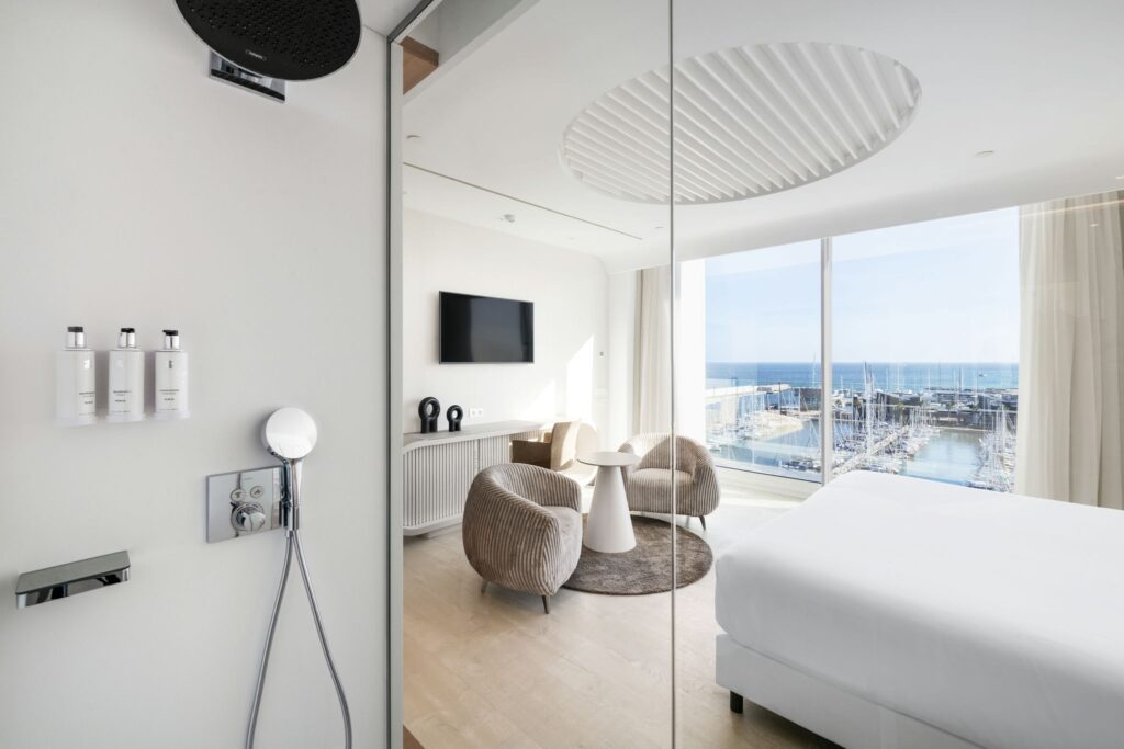 Habitación de Hotel Moderna y Lujosa con Vista al Puerto Una lujosa habitación de hotel moderna que exhibe grandes ventanales con una impresionante vista al puerto. La habitación cuenta con una cama blanca impecable, dos sillas de estilo y una pequeña mesa redonda para relajarse. Los accesorios de baño de cromo son visibles a la izquierda, equipados con comodidades contemporáneas de ducha. Un televisor de pared mejora la elegancia moderna del espacio, situado frente al acogedor área de estar.
