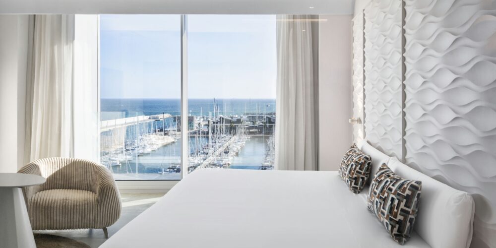 Una habitación de hotel moderna y espaciosa con una cama grande vestida con sábanas blancas y cojines decorativos, que cuenta con luz natural proveniente de una gran ventana que revela una pintoresca marina con barcos. El elegante diseño incluye muebles con estilo como un sillón y una mesa circular, acentuados por paneles de pared texturizados únicos.