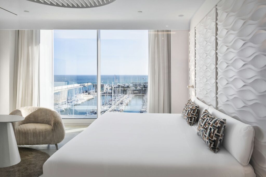 Habitación de Hotel Moderna y Lujosa con Vista a la Marina Una habitación de hotel moderna y espaciosa con una cama grande vestida con sábanas blancas y cojines decorativos, que cuenta con luz natural proveniente de una gran ventana que revela una pintoresca marina con barcos. El elegante diseño incluye muebles con estilo como un sillón y una mesa circular, acentuados por paneles de pared texturizados únicos.