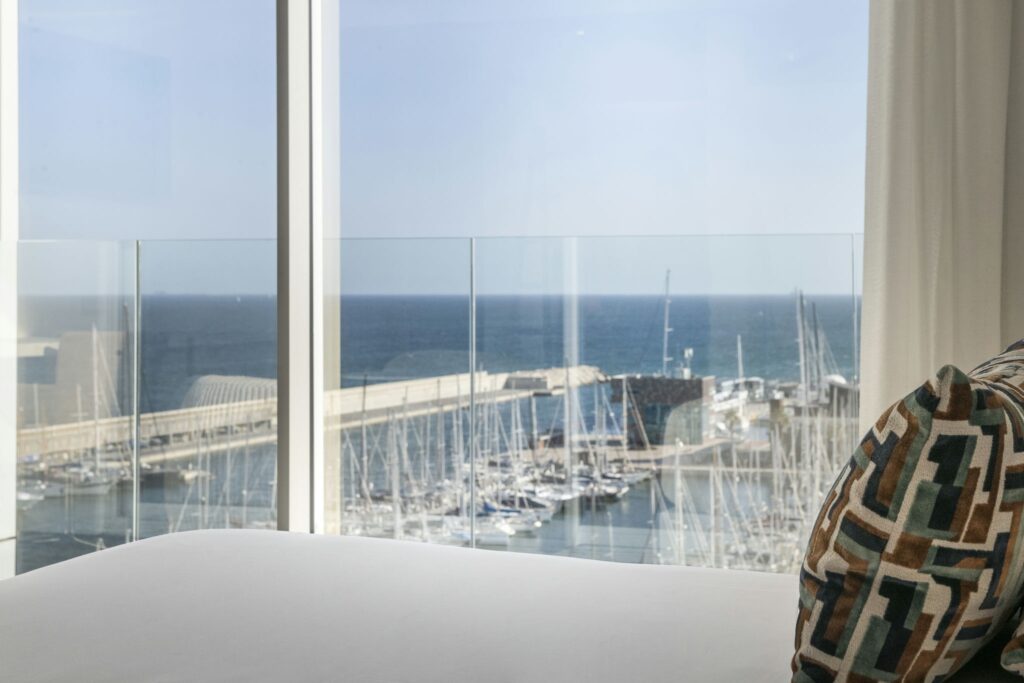 Vista Serene del Mar desde un Interior Moderno con Cojín Acolchado Un lujoso interior con grandes ventanales de cristal que ofrecen una impresionante vista del mar azul tranquilo y del cielo despejado, destacando una marina con yates y barcos blancos. Un cojín con patrón añade comodidad a la escena.