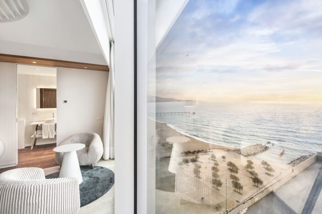 Habitación de Hotel Moderna con Vista Panorámica al Mar Una espaciosa habitación de hotel moderna con una vista panorámica del mar a través de grandes ventanas, sillas de tela acanalada elegantes y un ambiente tranquilo adecuado para la relajación.