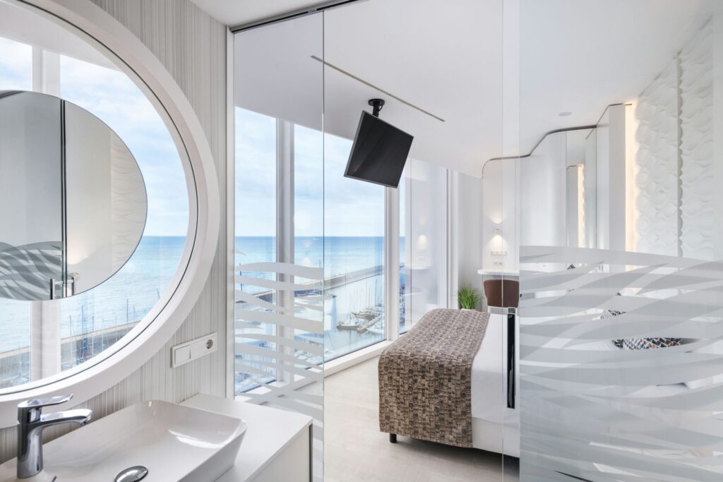 Habitación de hotel contemporánea con vistas al océano y diseño elegante Una habitación de hotel moderna con grandes ventanas de vidrio con vista al océano, un baño elegante con un espejo circular y particiones de vidrio, una cama cómoda con un cobertor estampado y luz natural inundando el espacio.