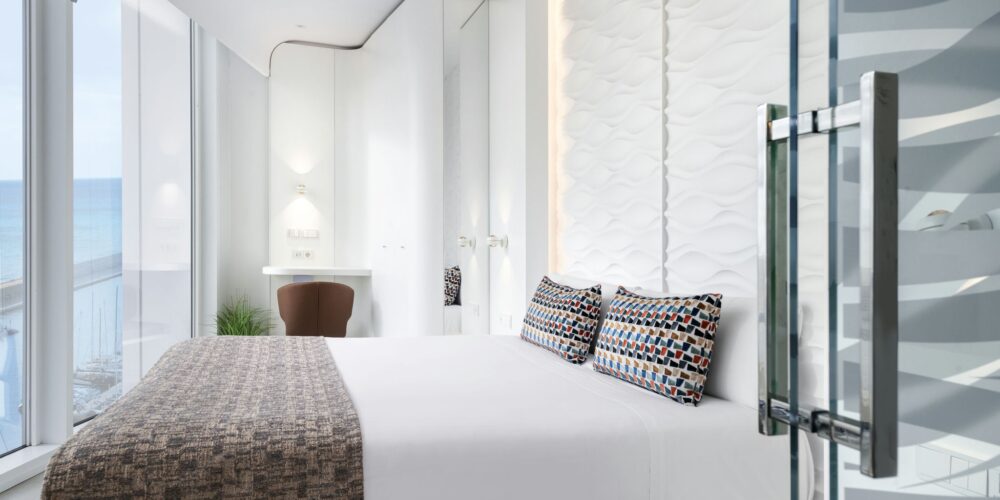 Una habitación de hotel moderna con grandes ventanas con vista al océano, un diseño minimalista, una cama blanca y acogedora con coloridos cojines decorativos, y un panel de vidrio que conduce al baño.