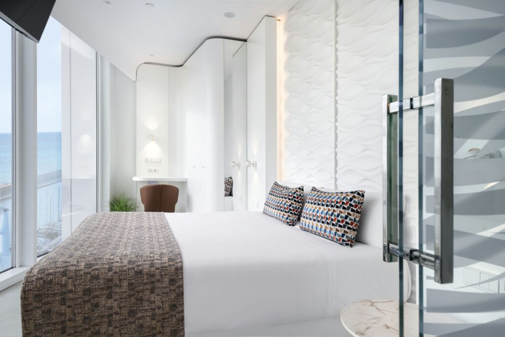 Habitación de Hotel Moderna y Elegante con Vista al Océano Una habitación de hotel moderna con grandes ventanas con vista al océano, un diseño minimalista, una cama blanca y acogedora con coloridos cojines decorativos, y un panel de vidrio que conduce al baño.