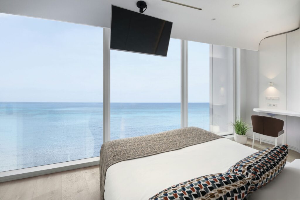 Habitación Moderna Elegante con Vista al Mar y Ambiente Acogedor Una habitación elegante que presenta una cama grande adornada con cojines decorativos, ventanas amplias que enmarcan una hermosa vista al océano, un televisor de pantalla plana en la pared y un pequeño espacio de trabajo. La habitación cuenta con paredes de color claro, piso de madera y toques de vegetación para una atmósfera serena.