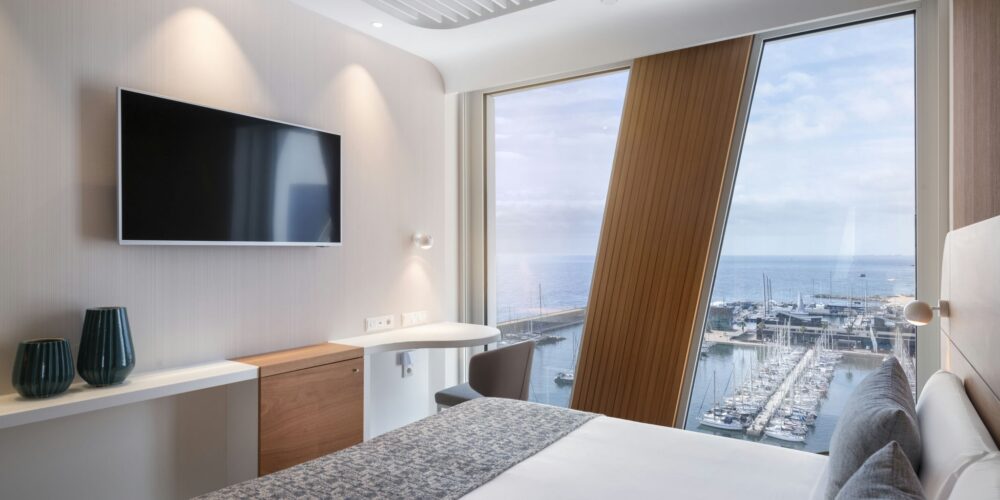 Habitación de hotel moderna que muestra una acogedora cama gris, un televisor de pantalla plana en la pared y un espacio de trabajo elegante con una silla. Las grandes ventanas revelan una impresionante vista del puerto y el océano con varios barcos, todo acentuado por la decoración neutral de la habitación y dos jarrones verdes decorativos.