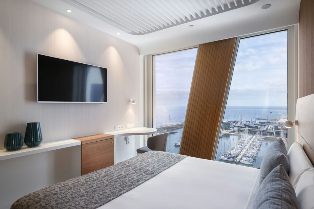 Habitación de hotel moderna y elegante con vista al puerto Habitación de hotel moderna que muestra una acogedora cama gris, un televisor de pantalla plana en la pared y un espacio de trabajo elegante con una silla. Las grandes ventanas revelan una impresionante vista del puerto y el océano con varios barcos, todo acentuado por la decoración neutral de la habitación y dos jarrones verdes decorativos.