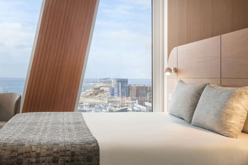 Habitación de Hotel Moderna con Vista Panorámica al Mar y al Paisaje Urbano Una habitación de hotel moderna y espaciosa con una gran cama con sábanas blancas y almohadas grises, una pared de madera cálida con lamas, y grandes ventanas que ofrecen una vista impresionante del mar y el horizonte de la ciudad.