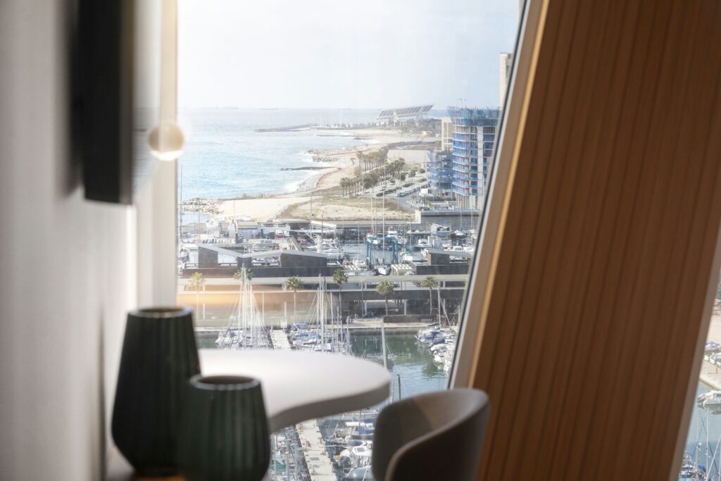 Vista Panorámica Costera desde un Espacio Interior Moderno Una escena interior contemporánea que muestra una gran ventana con una vista panorámica de un paisaje costero sereno que incluye una marina, barcos, una playa de arena y edificios modernos junto al agua, iluminados por una suave luz natural. En primer plano, lámparas elegantes y una mesa curva mejoran la atmósfera acogedora.
