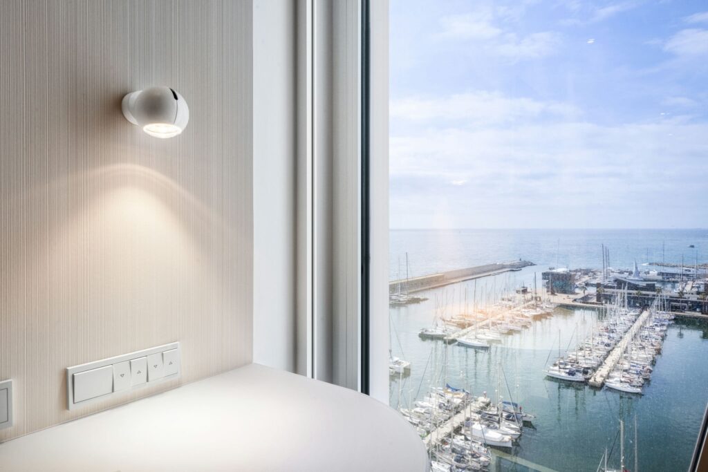 Diseño Interior Moderno con Lámpara Elegante y Vistas al Puerto Un interior moderno y elegante que muestra una lámpara redonda sobre una pared texturizada, con enchufes eléctricos visibles cerca. Detrás, grandes ventanas dan al sereno puerto lleno de yates bajo un cielo parcialmente nublado, permitiendo que la luz natural inunde el espacio y resalte su estética contemporánea.