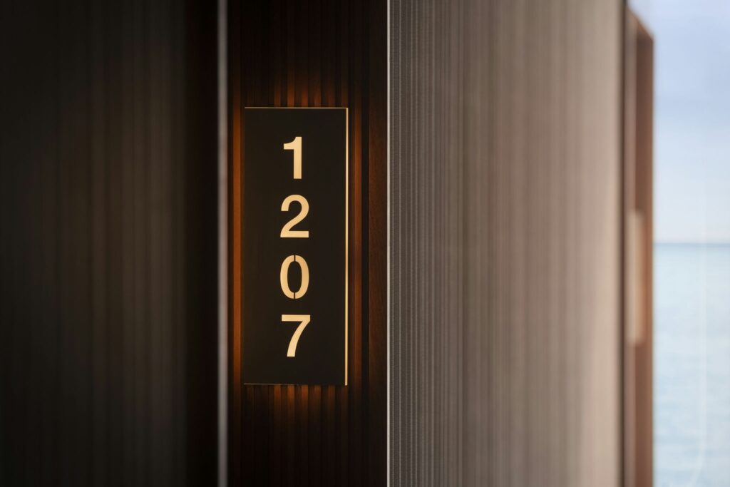 Puerta de hotel elegante con número de habitación iluminado 1207 Primer plano de una puerta de hotel que presenta una moderna placa de número de habitación iluminada que dice '1207', colocada contra un panel de madera texturizado, con un paisaje sereno insinuándose en el fondo.