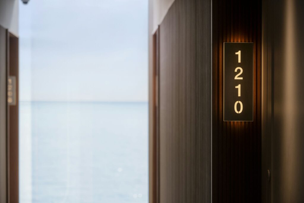 Elegante Pasillo de Hotel Costero con Número de Puerta 1210 Una vista cercana de un letrero de número de puerta que dice '1210' en un pasillo de hotel suavemente iluminado. El fondo presenta una imagen borrosa de agua, indicando la proximidad de un océano o lago, con paneles de madera elegantes a lo largo de las paredes.