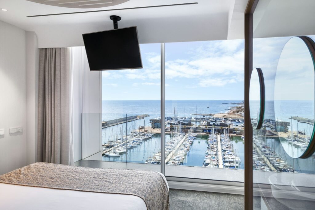 Habitación de Hotel Moderna y Lujosa con Vista a la Marina Amplia habitación de hotel con una cama grande y una manta texturizada, una pared de vidrio que exhibe una marina con botes y yates, y cortinas ligeras que realzan el ambiente brillante y aireado.