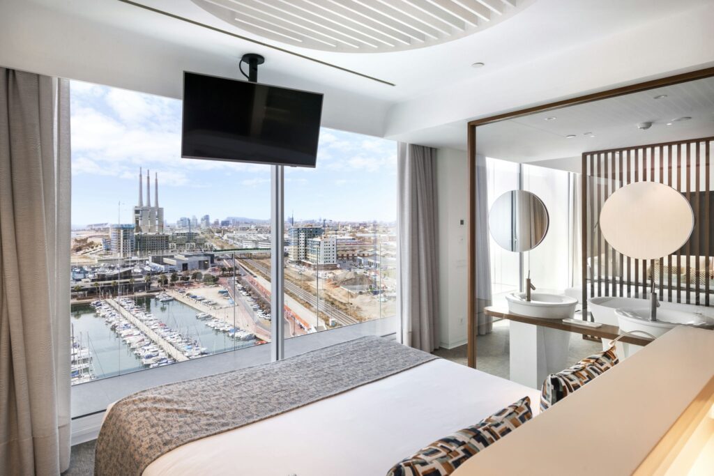 Habitación de hotel moderna con vista a la ciudad frente al mar Habitación de hotel elegante con grandes ventanas que ofrecen vista a la ciudad frente al mar, una cama bien hecha con una manta texturizada y almohadas decorativas, un televisor montado en la pared y un baño contemporáneo con dos espejos redondos y lavabos, todo diseñado en un estilo minimalista y elegante.