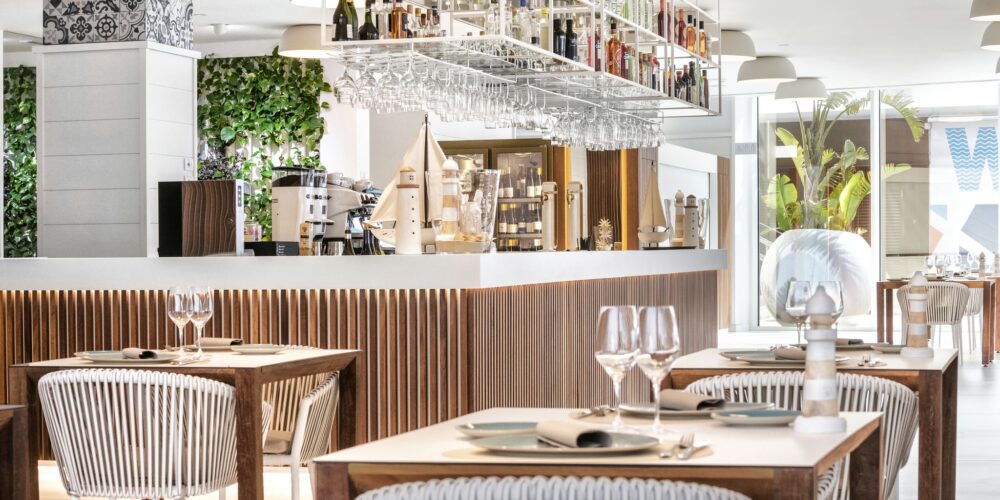 Interior de un restaurante moderno con mesas de madera preparadas con platos blancos y copas de vino, barra minimalista con varias botellas, estantes de vidrio en el techo, lámparas elegantes y abundante vegetación. El espacio es luminoso y aireado con grandes ventanales.