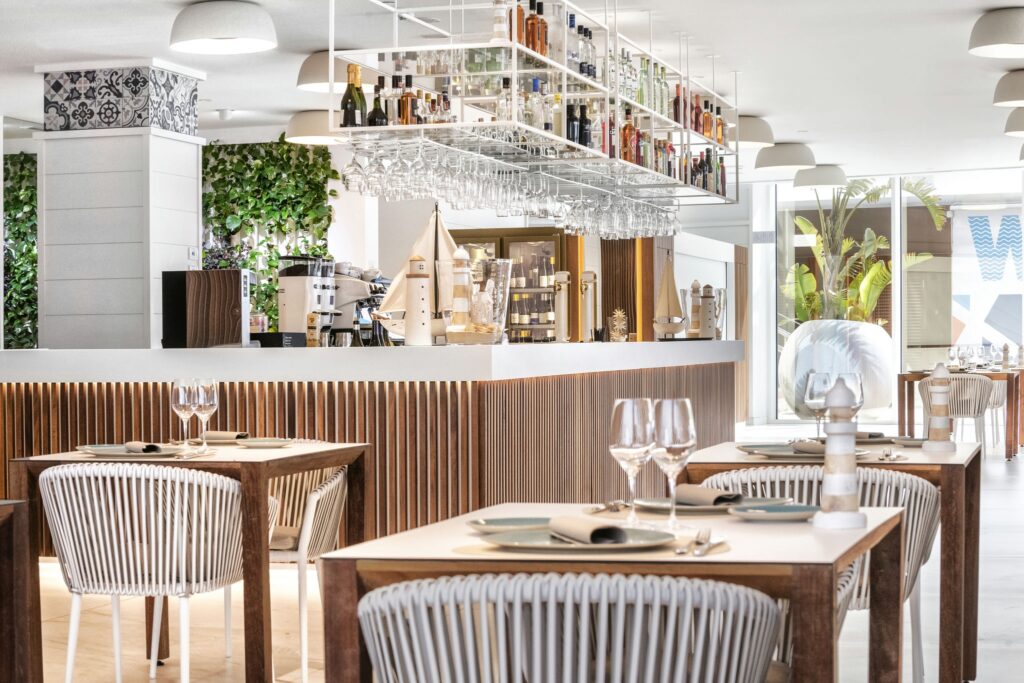 Interior Elegante y Moderno de Restaurante con Luz Natural Interior de un restaurante moderno con mesas de madera preparadas con platos blancos y copas de vino, barra minimalista con varias botellas, estantes de vidrio en el techo, lámparas elegantes y abundante vegetación. El espacio es luminoso y aireado con grandes ventanales.