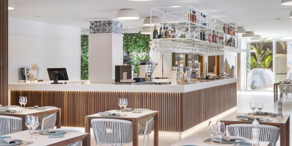 Un interior de restaurante moderno, luminoso y aireado que presenta un área de bar esbelta con una encimera blanca, acentos de madera y botellas colgantes. Mesas ordenadamente dispuestas con sillas de rayas grises están puestas con platos azules y cristalería, iluminadas por luces colgantes y complementadas con vegetación. Grandes ventanales permiten que la luz natural mejore la atmósfera acogedora para los comensales.