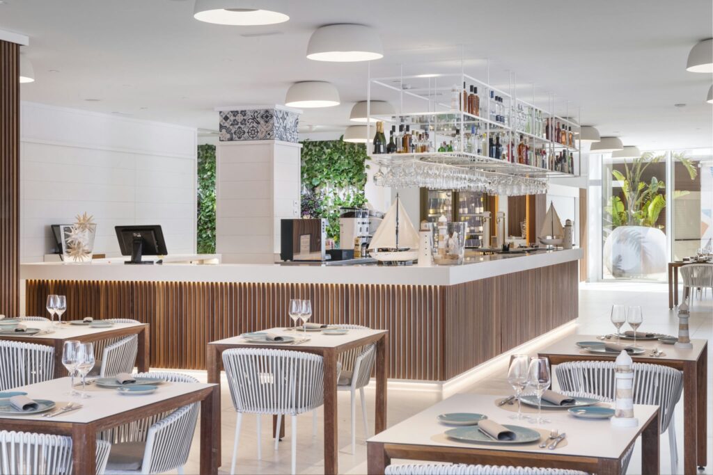 Interiores de Restaurante Moderno y Elegante con Área de Bar Esbelta Un interior de restaurante moderno, luminoso y aireado que presenta un área de bar esbelta con una encimera blanca, acentos de madera y botellas colgantes. Mesas ordenadamente dispuestas con sillas de rayas grises están puestas con platos azules y cristalería, iluminadas por luces colgantes y complementadas con vegetación. Grandes ventanales permiten que la luz natural mejore la atmósfera acogedora para los comensales.