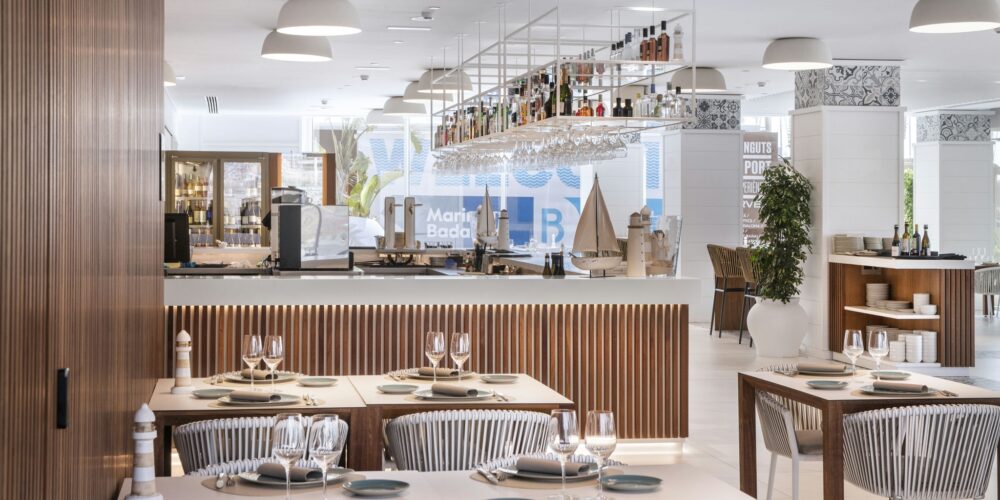 Un interior de restaurante moderno que muestra un diseño elegante, con elementos de madera, una paleta de colores claros y mesas de comedor bien dispuestas con platos blancos y cristalería, complementadas por muebles elegantes y vegetación decorativa.