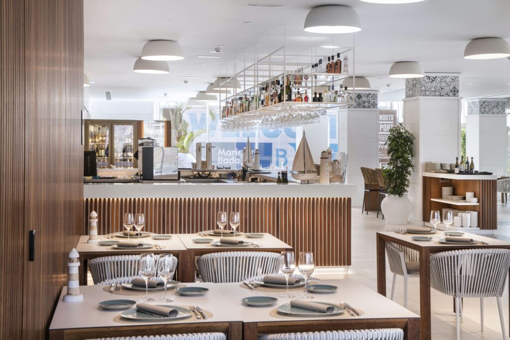 Interiores de Restaurante Moderno y Elegante con Acabados de Madera y Diseño Contemporáneo Un interior de restaurante moderno que muestra un diseño elegante, con elementos de madera, una paleta de colores claros y mesas de comedor bien dispuestas con platos blancos y cristalería, complementadas por muebles elegantes y vegetación decorativa.