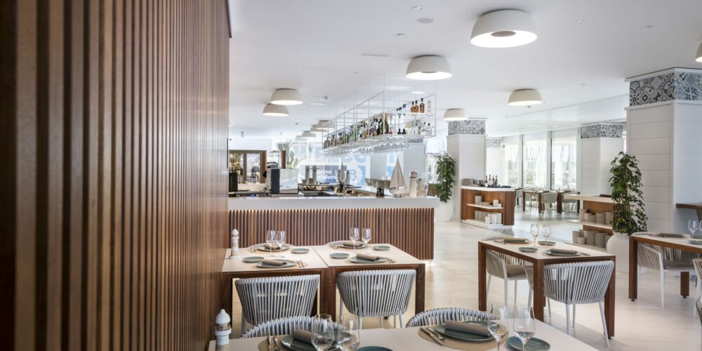 Un interior de restaurante moderno y elegante que presenta mesas organizadas ordenadamente con manteles blancos y platos de acento azules, sillas con respaldo acanalado de estilo, un área de bar con diversas botellas y una iluminación brillante y difusa. Plantas decorativas y azulejos estampados realzan la atmósfera acogedora.