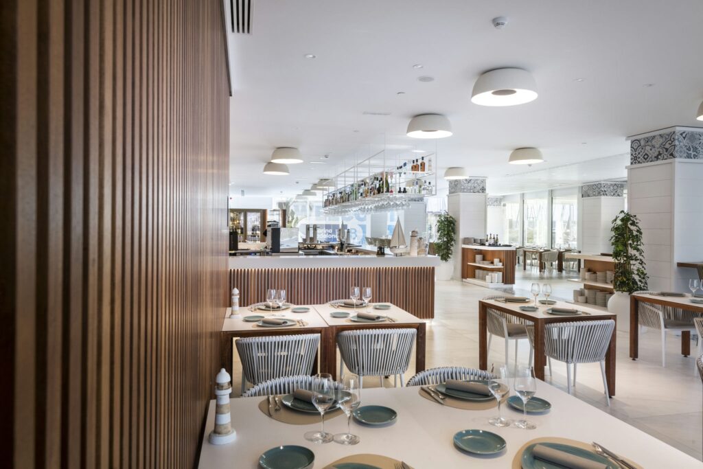 Interior de Restaurante Moderno y Elegante con Diseño Espacioso Un interior de restaurante moderno y elegante que presenta mesas organizadas ordenadamente con manteles blancos y platos de acento azules, sillas con respaldo acanalado de estilo, un área de bar con diversas botellas y una iluminación brillante y difusa. Plantas decorativas y azulejos estampados realzan la atmósfera acogedora.