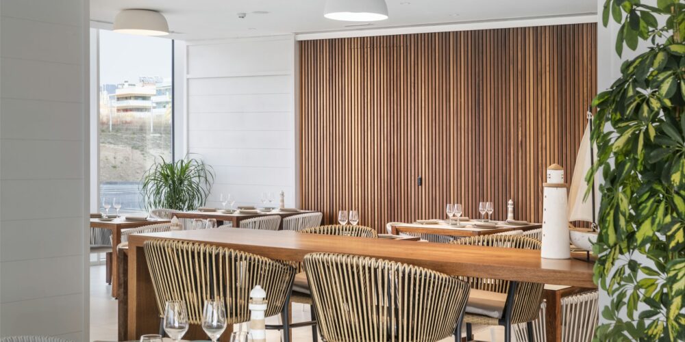 Vista interior de un restaurante moderno que muestra un diseño espacioso con decoración de madera, mesas elegantemente arregladas con cristalería, sillas de rayas elegantes y grandes ventanas que permiten la entrada de luz natural. Una atmósfera contemporánea complementada por listones verticales de madera en las paredes y un acento de planta.