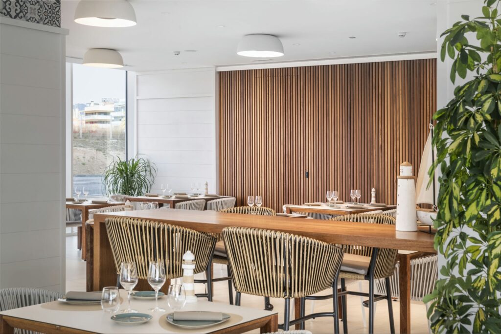 Interior de Restaurante Moderno con Mesas Elegentemente Puestas y Detalles de Madera Vista interior de un restaurante moderno que muestra un diseño espacioso con decoración de madera, mesas elegantemente arregladas con cristalería, sillas de rayas elegantes y grandes ventanas que permiten la entrada de luz natural. Una atmósfera contemporánea complementada por listones verticales de madera en las paredes y un acento de planta.