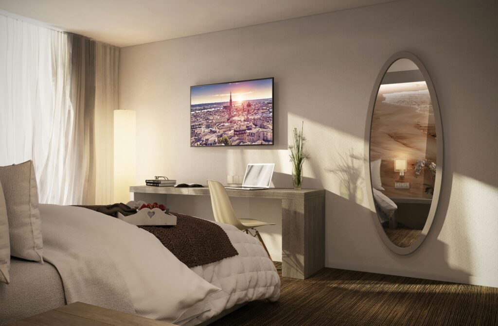 Diseño de habitación moderna y elegante en Barcelona - Proyectos de interiorismo de alta gama Habitación moderna y elegante en Barcelona con cama grande, escritorio contemporáneo, planta decorativa y espejo ovalado. La luz natural entra a través de cortinas transparentes, creando un ambiente luminoso que destaca la funcionalidad y los materiales de alta calidad como madera y vidrio.
